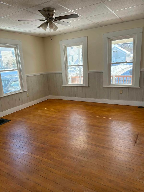 Empty room with painted walls and wainscot, white window trim, a ceiling fan and a hardwood floor