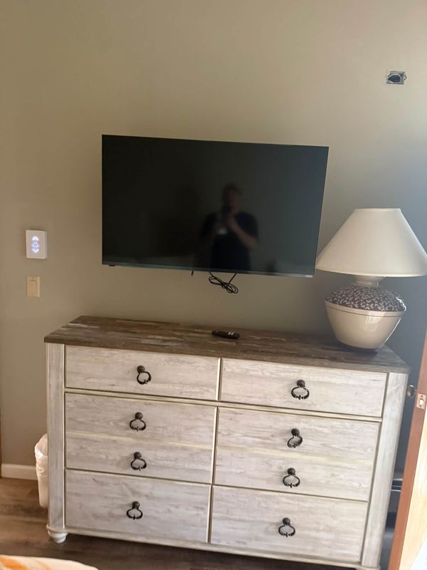 Television mounted flat on the wall above a white dresser with a lamp beside it