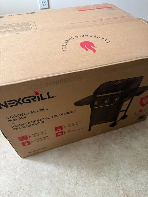 Boxed three burner gas grill sitting on a vinyl floor, not yet assembled