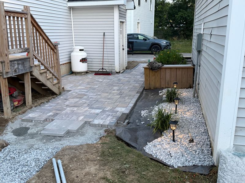 Paver patio laid out beside the house with a planted raised bed and path lighting