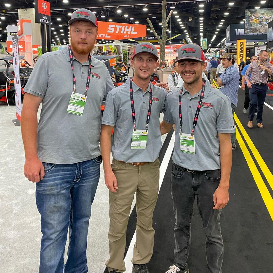 Three Buckeye Lawns crew members in gray company polos and red and gray caps standing together at a lawn and landscape trade show