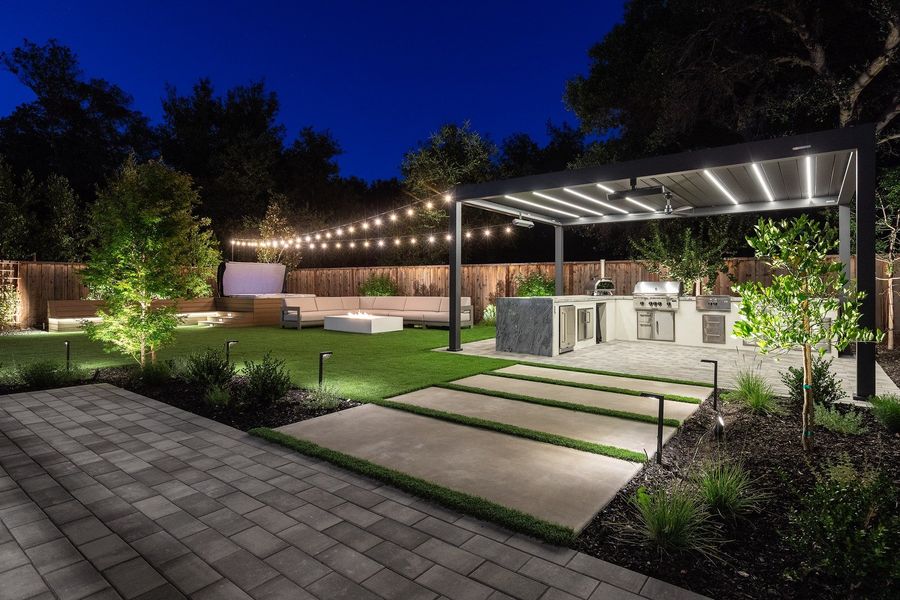 Grey paver terrace and concrete stepping pads at dusk with a lit pergola, kitchen island and path lights beyond