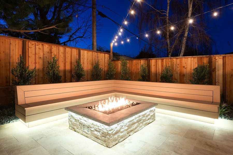 Night view of a lit gas fire feature clad in stacked stone inside an L-shaped bench with underglow, against a cedar fence