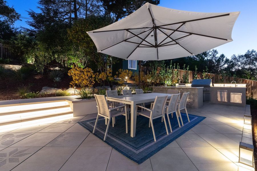 Night view of the dining table under a white umbrella with the kitchen island glowing beside a lit stepped planter