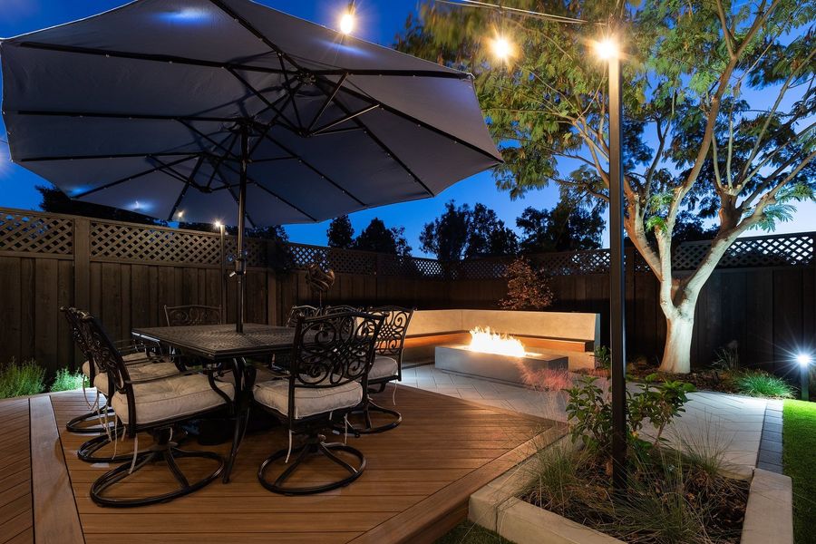 Dusk view of a dining table under a large umbrella on a wood-look deck with string lights and a glowing fire table behind