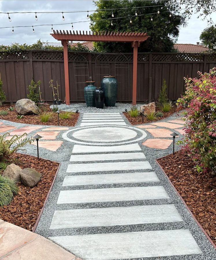 Path of pale concrete strip pavers set in gravel, with a circular medallion, flagstone edges, mulch beds and a small pergola framing large teal and black ceramic jars.