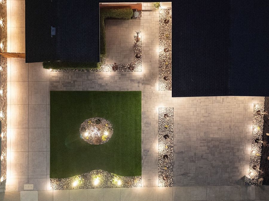 Night aerial of the same turf square and paver court with lights glowing along the paving edges and in the beds