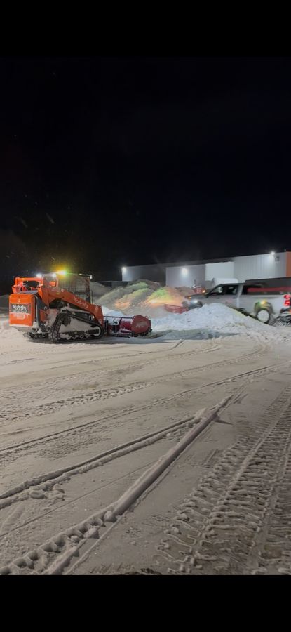 Orange loader with its lights on pushing snow across a paved area outside a building at night