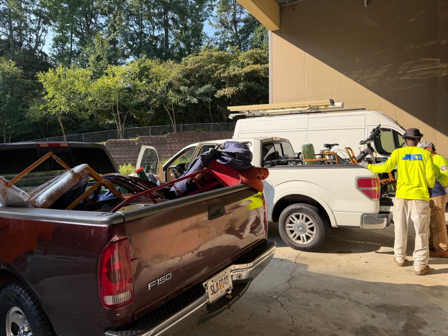 Crew members in yellow safety shirts beside a pickup truck loaded with furniture and a white truck