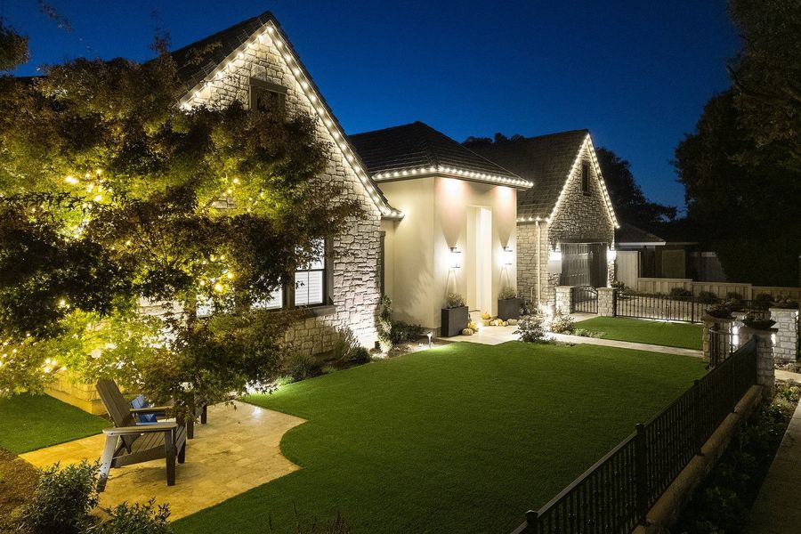 Night view of a lit lawn, black iron fence with stone pillar lights and a stone gable under a dark blue sky