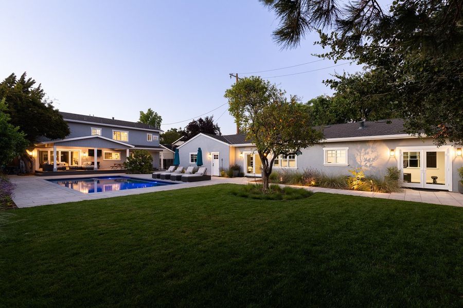 Dusk view across a green lawn with a citrus tree in a grass bed, slab paving along the far edge and lit wall lanterns on the single-story wing