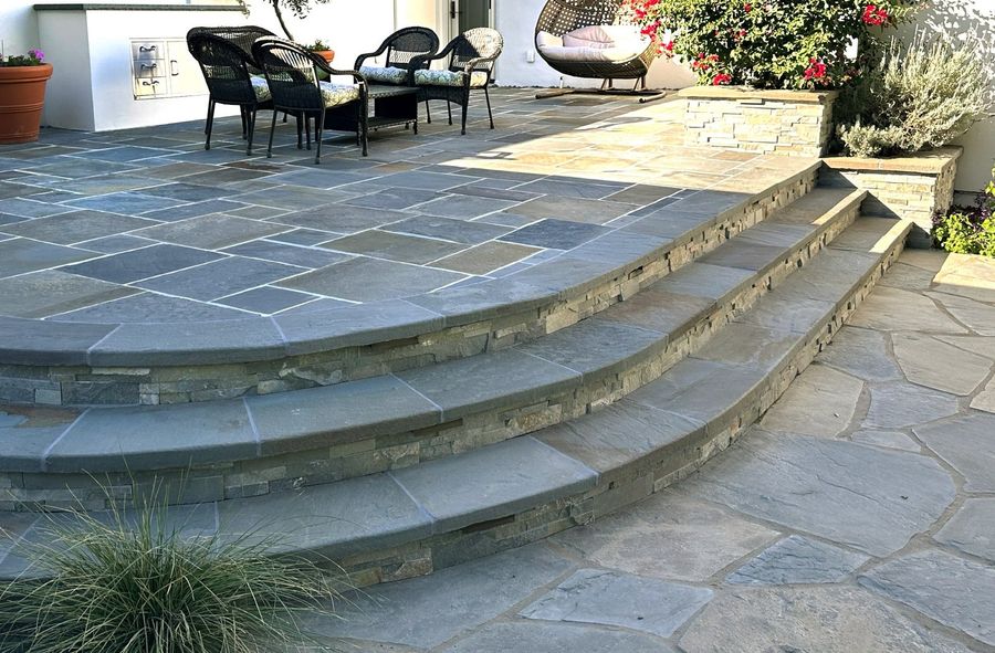 Three curved bluestone steps faced with stacked stone rising to a cut-stone patio with wicker seating, a stone planter with bougainvillea and lavender, and irregular flagstone paving at the bottom.