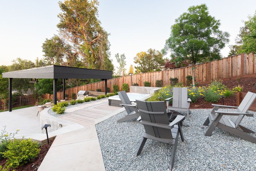 Four gray Adirondack chairs around a fire bowl on gravel, with yellow flowering perennials and the pavilion below