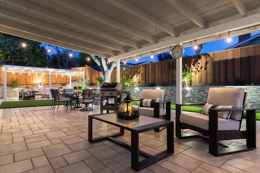 Covered patio at dusk with lounge chairs and a coffee table on pavers, string lights overhead and a stone wall beyond