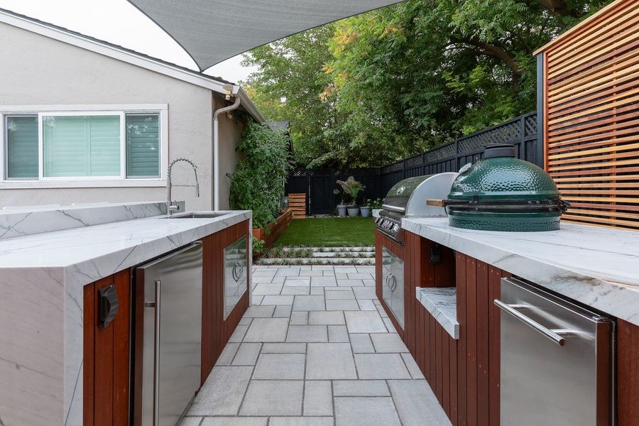 Stone countertop with sink, stainless fridge and drawers, grill and green ceramic cooker, with the lawn and stepping stones beyond