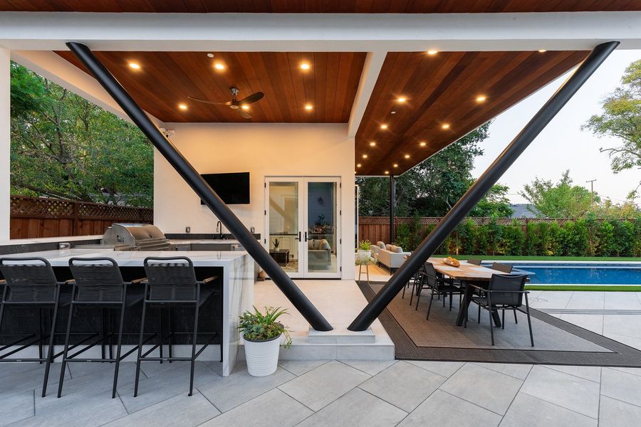 Crossed dark steel struts holding a timber-lined ceiling with recessed lights, over a bar counter with stools and a dining table