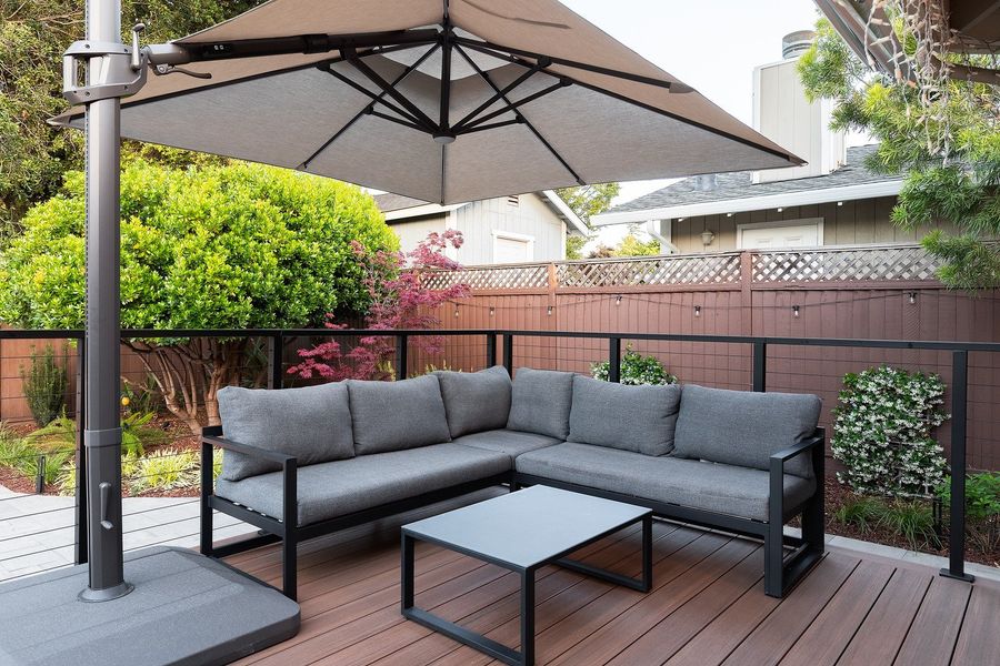 Outdoor sectional and coffee table on the deck beneath a taupe umbrella, with a Japanese maple behind the railing