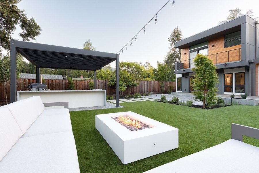 Rectangular white concrete fire table alight between two white sofas on turf, with the louvered pergola and modern house behind