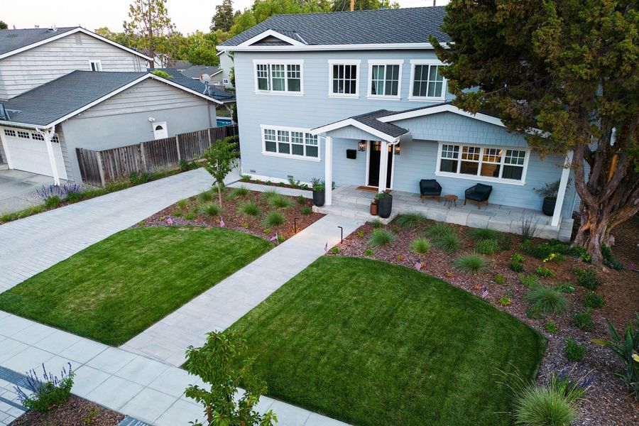 Elevated view of a two-story blue house with a slab walk crossing two lawns to a covered front porch, with grass and shrub beds beside it