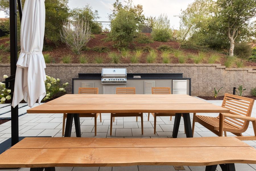 Teak dining table in front of a built-in grill island set against a stepped stone retaining wall and a planted hillside