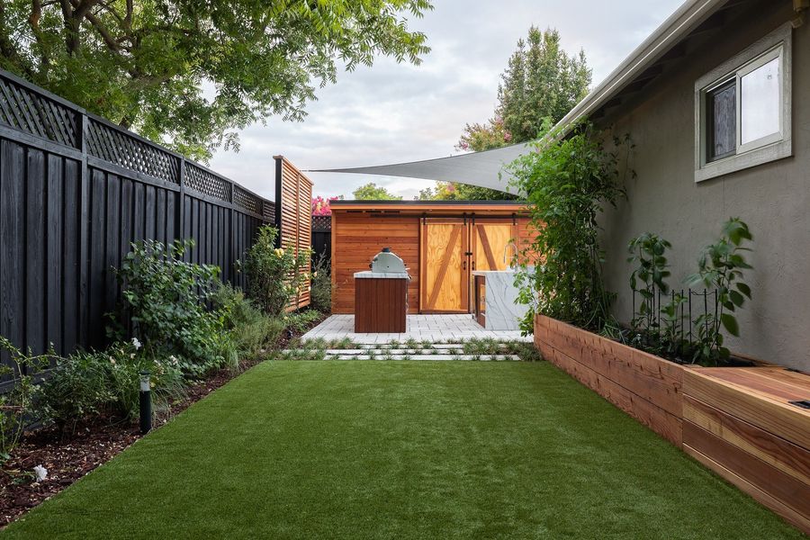 Lawn bordered by layered planting and a raised cedar planter, leading to a cedar outdoor kitchen under a shade sail