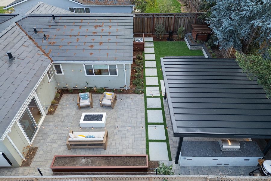 Aerial view of the paver court with fire table and seating, a path of large pavers set in turf, a raised planter and the pavilion roof