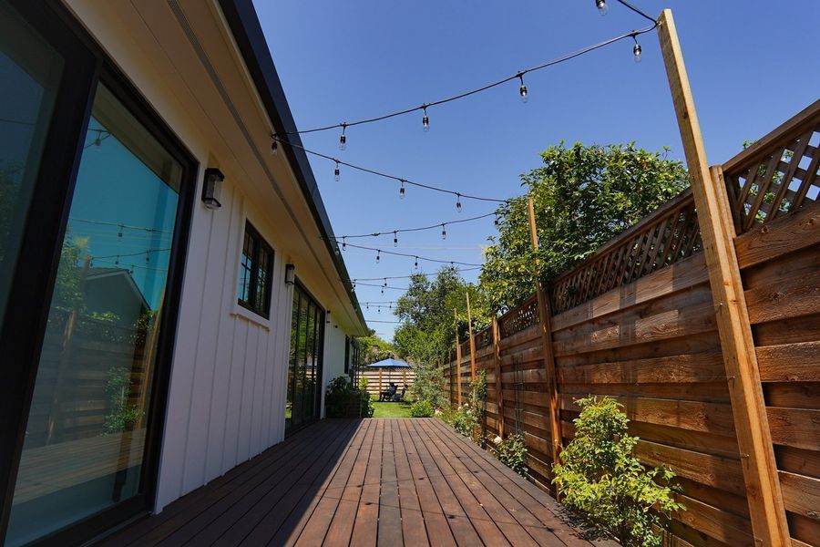 Narrow wood deck walkway running along a white house beside a tall horizontal-slat wood fence, with string lights overhead and shrubs along the fence.