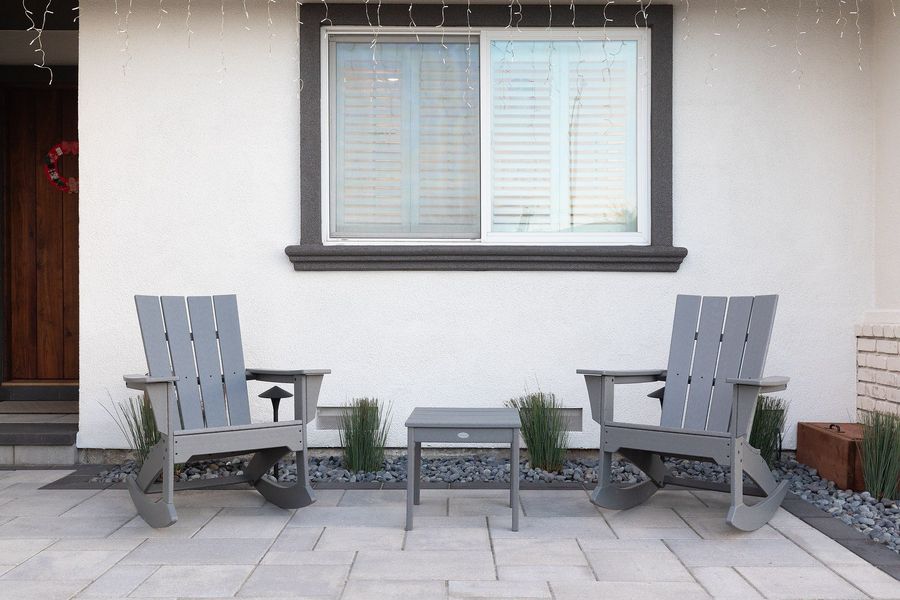 Daytime view of two gray Adirondack rocking chairs and a side table on a paver porch with river rock and grasses