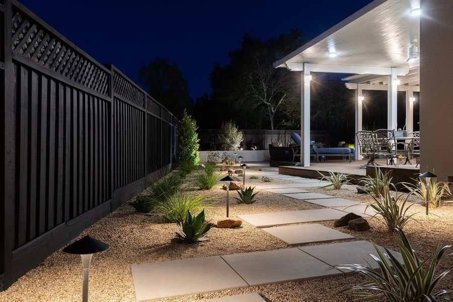 Night view along a dark fence of square pale slabs through a lit gravel bed of agave and grasses, with the covered patio ahead