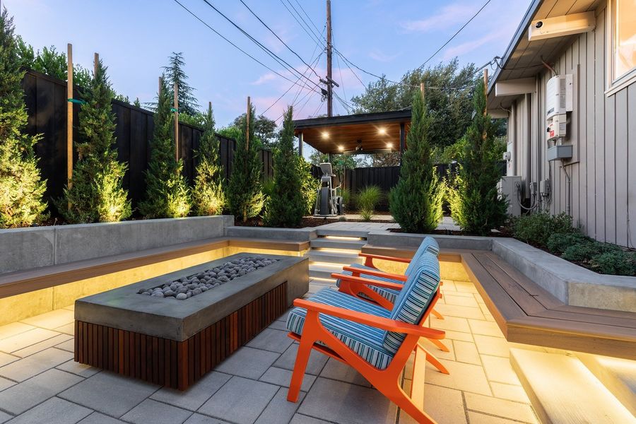 Fire table and built-in benches with glowing under-edge lighting, tall conifers and a wood pavilion at the far end
