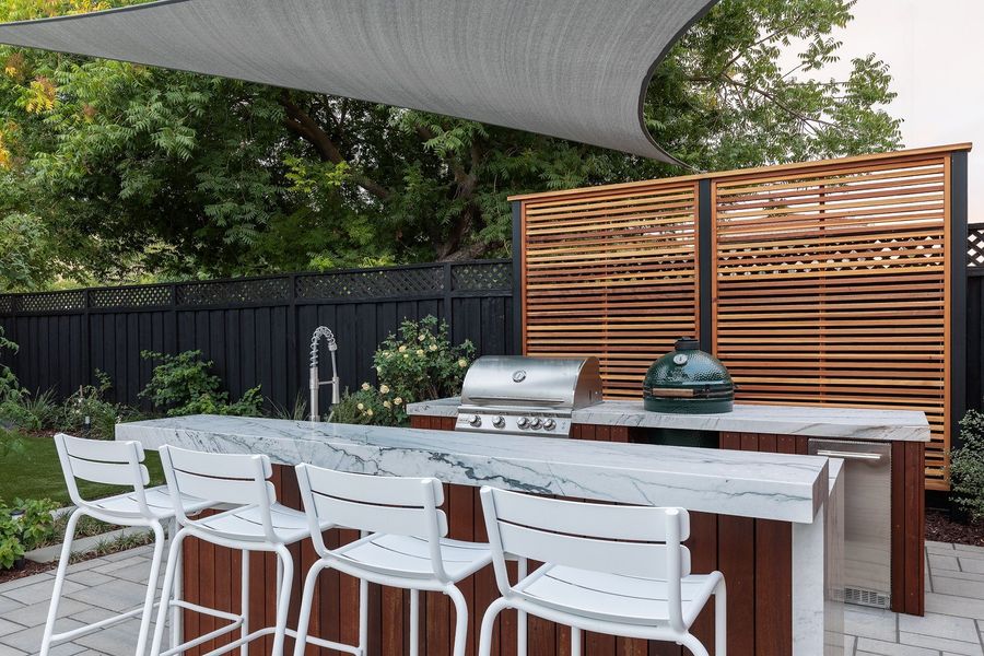 Daytime view of the stone bar with white stools, sink, grill and ceramic cooker beside a cedar screen
