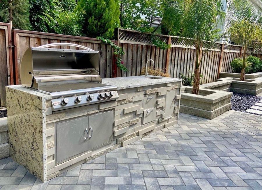 Built-in outdoor kitchen with a stainless gas grill open, a granite counter with a sink and tall faucet, stacked stone veneer on the island, and stainless doors and drawers, on a gray herringbone paver patio beside a stacked-block planter.