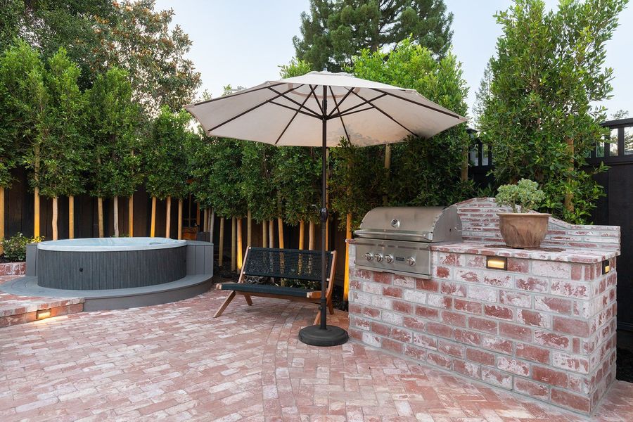 Brick grill island with a stainless grill, bench and umbrella on herringbone brick, with a tall evergreen screen behind