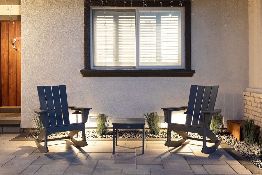 Dusk view of two dark Adirondack chairs and a side table on pavers, with river rock, grasses and uplighting under a lit window