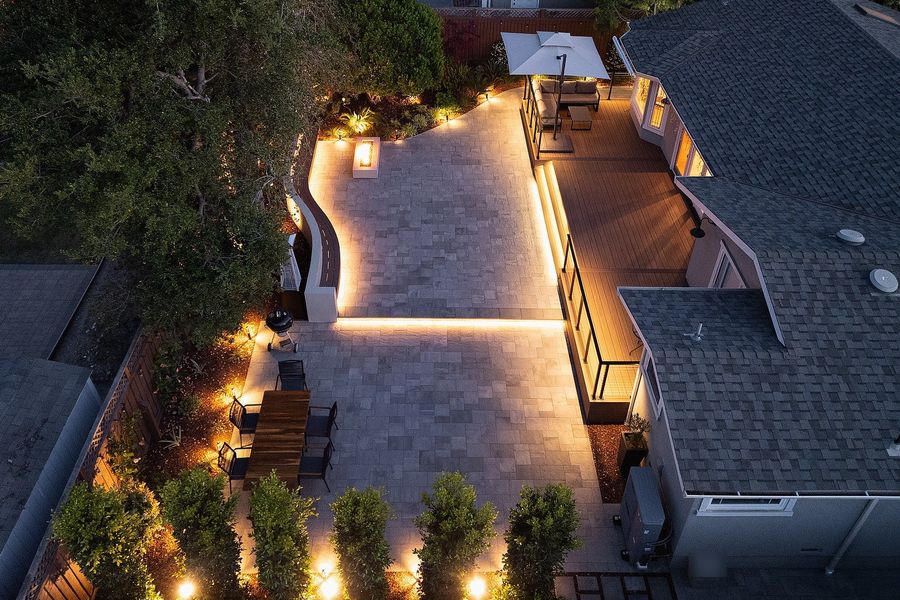 Night aerial of the paver terrace outlined in warm light, with the lit deck and uplit hedges