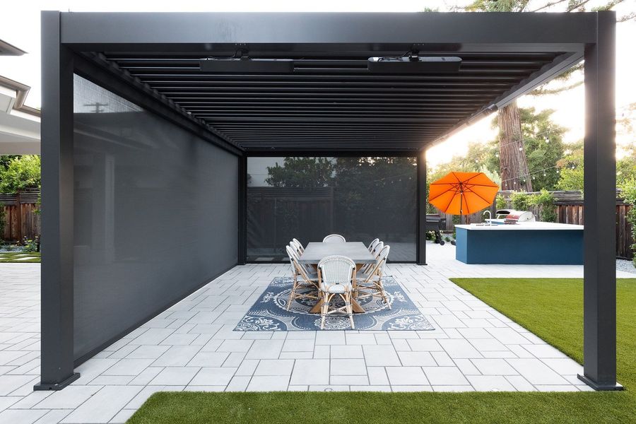 Pergola with a grey screen lowered along one side over a dining table and rug, with the blue kitchen island beyond