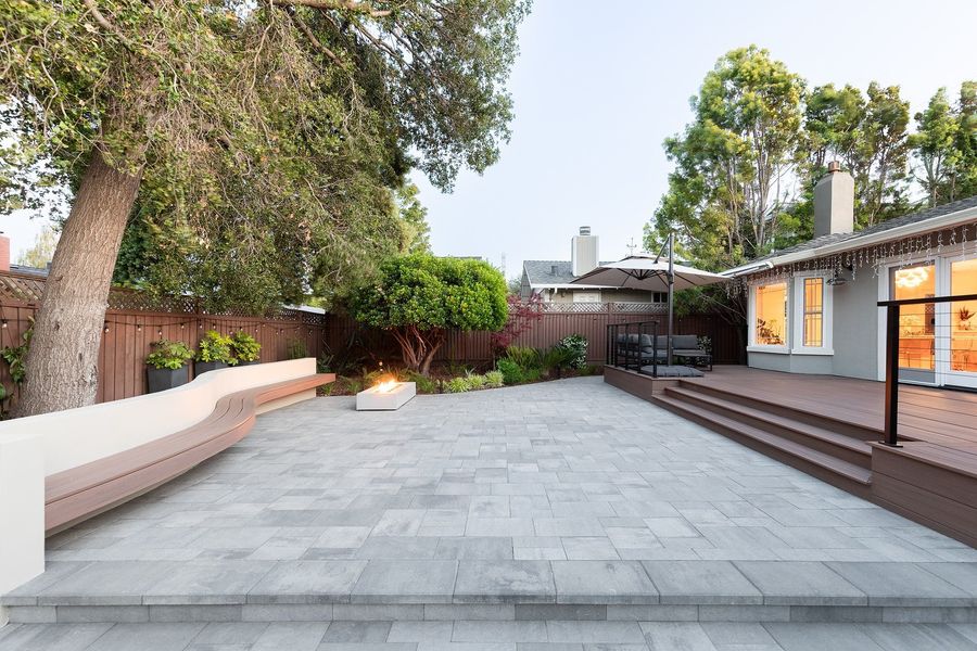Daytime view across a gray multi-tone paver court to a stepped wood-look deck and a curved bench with a fire table