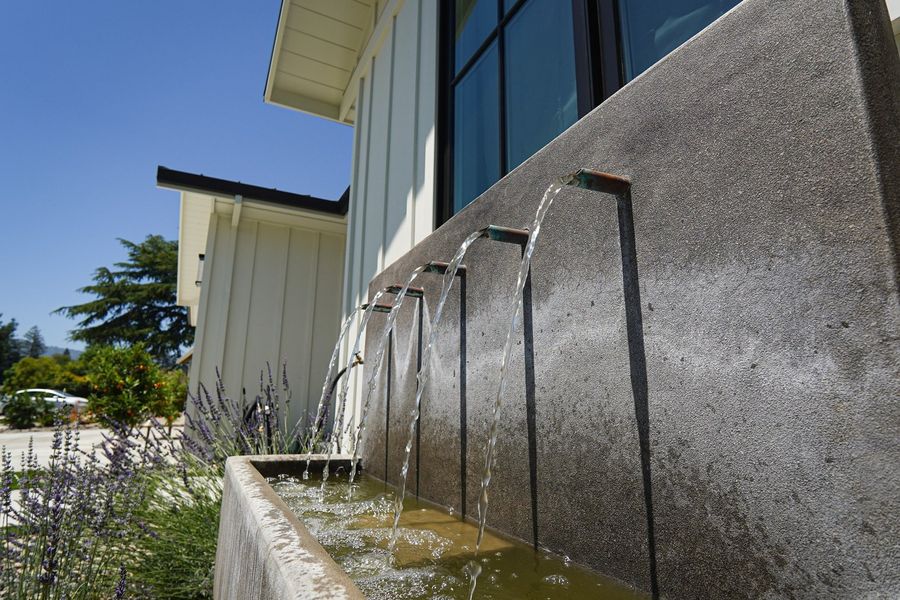 Several thin jets of water falling from a spouted wall into a long concrete trough against a white house, with lavender and gravel in front.