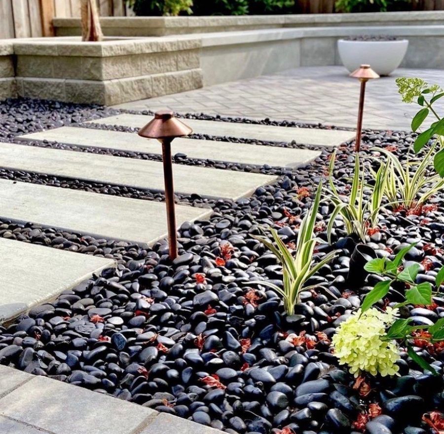 Close view of pale rectangular stepping-stone slabs set in black polished pebbles beside a copper path light, with variegated grass, a hydrangea bloom, and a capped stacked-block wall and white fire bowl behind.