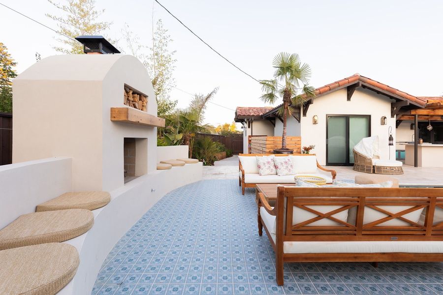 Daytime view of a stucco fireplace with a curved seat wall, blue patterned tile floor and teak daybeds, the white house beyond