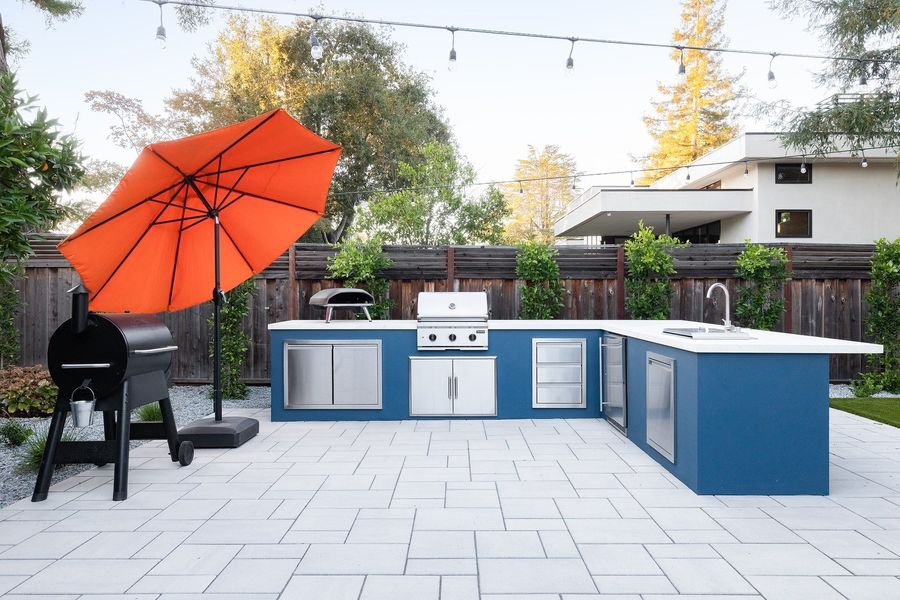 L-shaped blue kitchen island with a built-in grill, sink, stainless drawers and a pizza oven under an orange umbrella