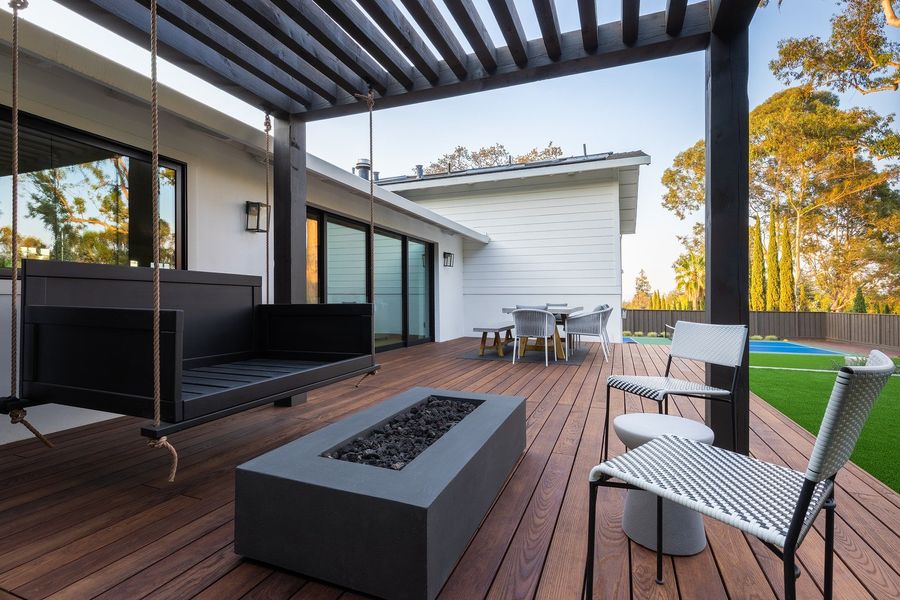 Hardwood deck with a concrete fire table, woven chairs and a hanging swing under a black pergola in warm afternoon light