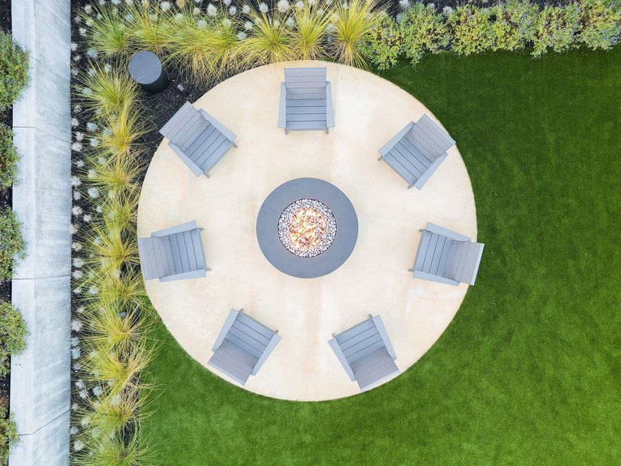 Straight-down aerial of a round concrete patio with a fire bowl and eight chairs, ornamental grasses and lawn