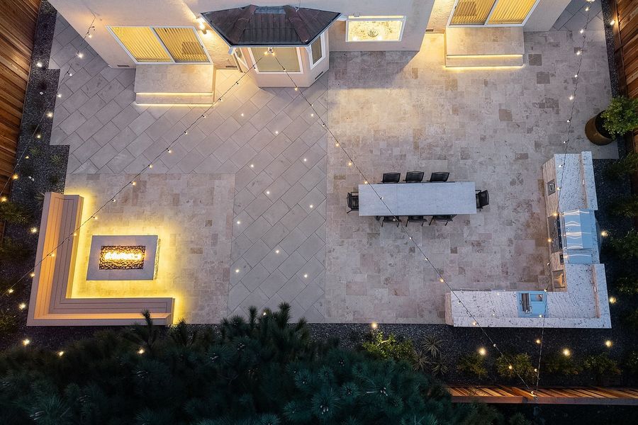Aerial night view of the paved patio with the fire bench, dining table and kitchen island and string lights crossing above