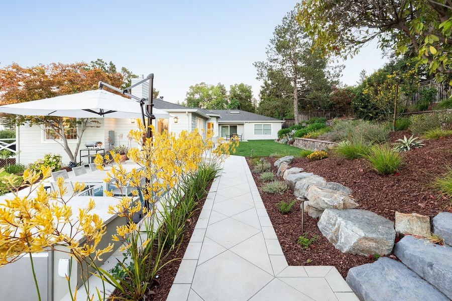 Pale paved path with angled joints running past yellow kangaroo paw, grasses and large blue-grey boulders in mulched beds