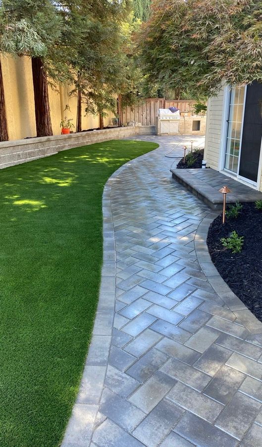 Curving gray herringbone paver path with a border course, a paver step up to a sliding glass door, copper path lights in a black mulch bed, and the outdoor kitchen visible at the far end.