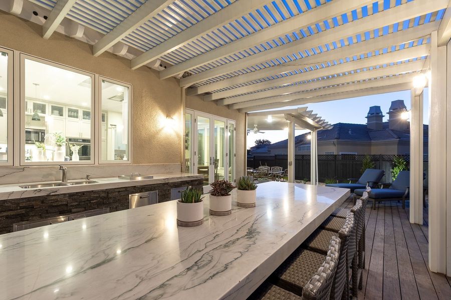 Long veined white counter running under the pergola at dusk, with the kitchen window at left and lounge chairs beyond