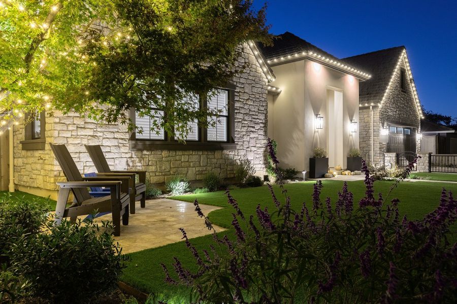Close twilight view of two lounge chairs on a travertine patio with purple salvia in the foreground and lit stucco entry behind