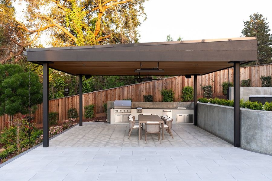 Flat-roofed pavilion with a wood ceiling over a stainless outdoor kitchen, a long dining table and patterned floor tile