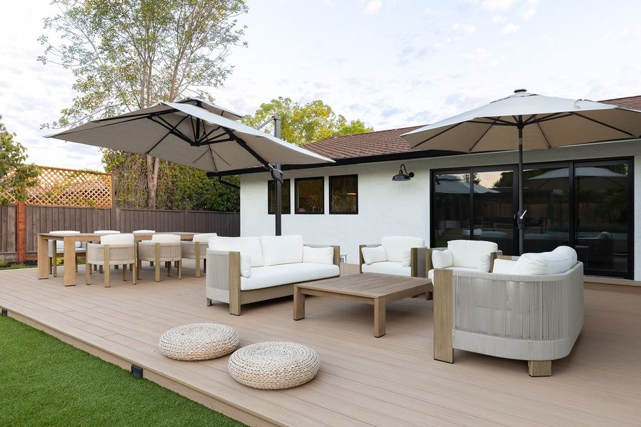 Light composite deck beside the house with a dining table, white sofas, two large cantilever umbrellas and black sliding doors